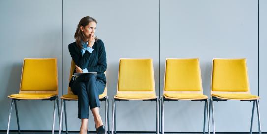 Nachdenklich wirkende Frau im Business-Outfit sitzt allein auf einer gelben Stuhlreihe in einem Wartebereich und blickt zur Seite