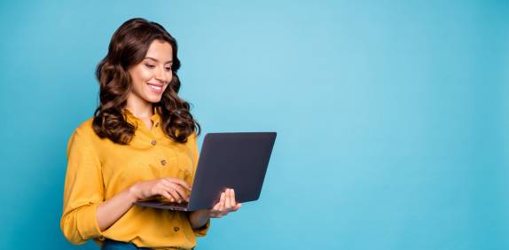 Junge Frau mit gewellten braunen Haaren trägt eine gelbe Bluse und blaue Hose, steht vor einem blauen Hintergrund und arbeitet lächelnd an einem Laptop
