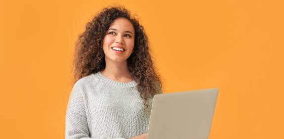 Junge Frau mit lockigem braunem Haar trägt einen hellgrauen Strickpullover, steht vor einem orangefarbenen Hintergrund und lächelt, während sie einen Laptop in den Händen hält
