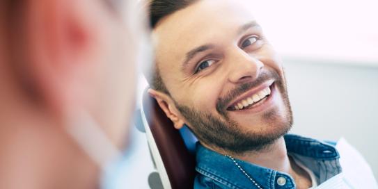 Junger Mann sitzt entspannt auf einem Behandlungsstuhl beim Zahnarzt und lächelt zufrieden in Richtung der behandelnden Person