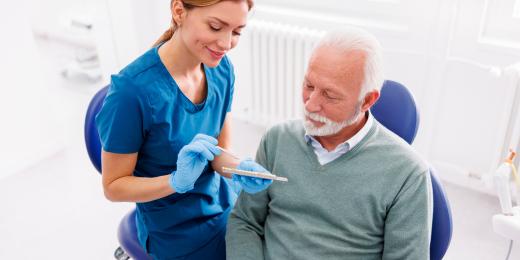 Zahnärztin in blauer Kleidung erklärt einem älteren männlichen Patienten mit grauem Haar ein Zahnmodell